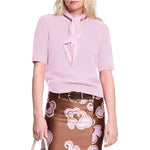 A woman wears the Dorothee Schumacher Fluffy Statements Pullover, a soft knit light pink short sleeve sweater, paired with a matching scarf and brown skirt with pink and white swirls, holding a white bag.