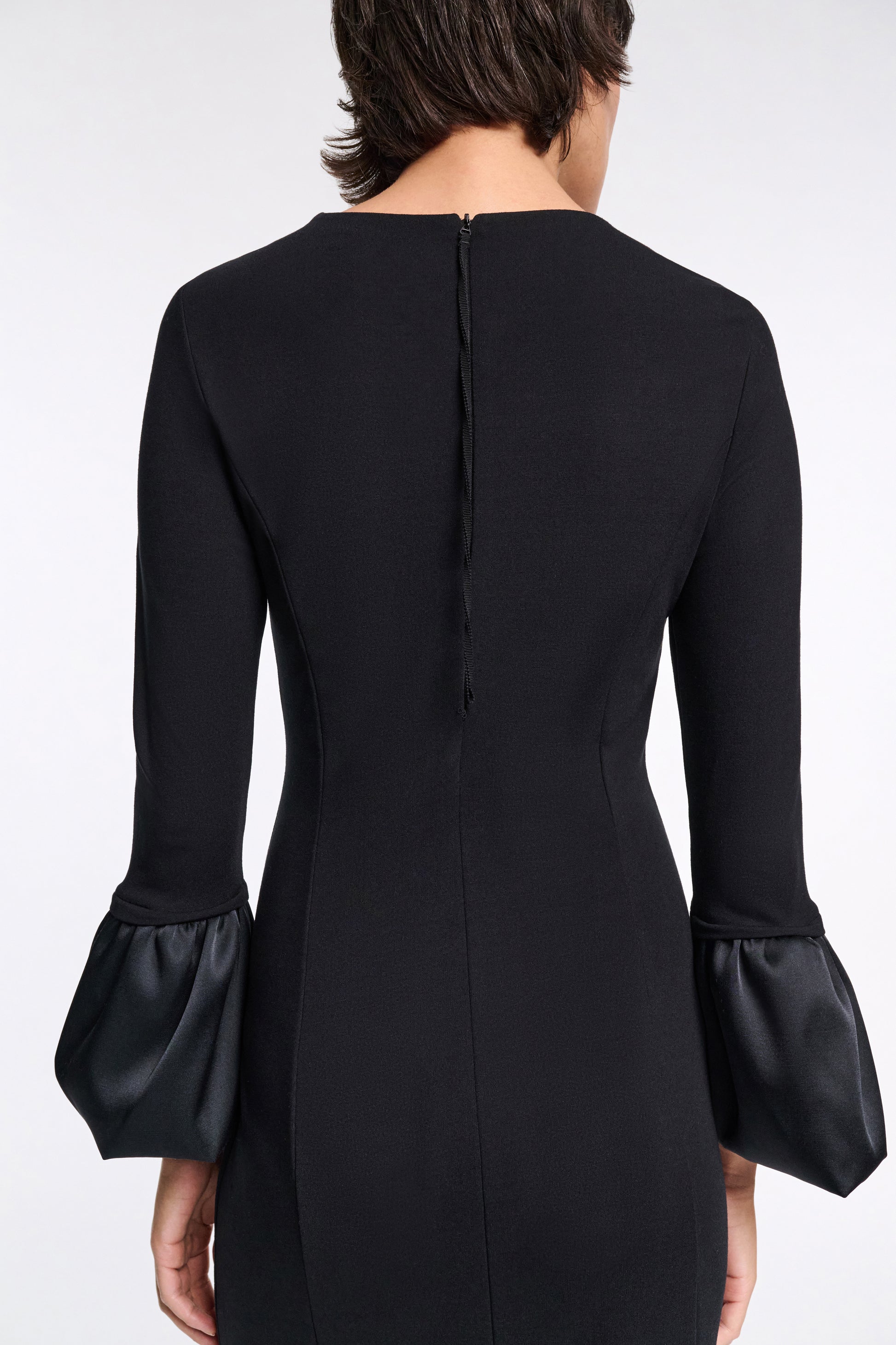 A person is shown from the back wearing the Dorothee Schumacher Cloudy Allure Dress, a fitted black midi dress with long sleeves and satin bubble cuffs, set against a plain background.
