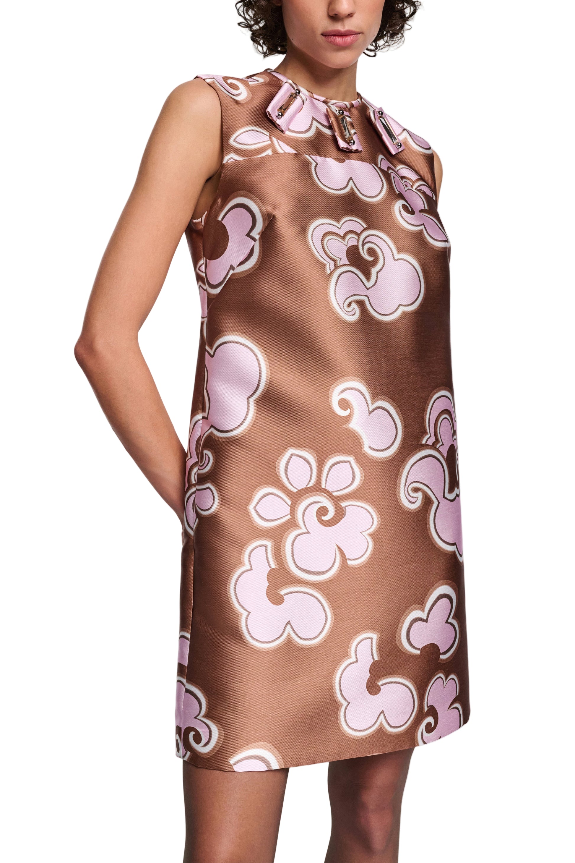 A person wears the Dorothee Schumacher Floral Statement Dress, a sleeveless brown satin shift featuring pink and white abstract floral patterns and a bow at the neckline.