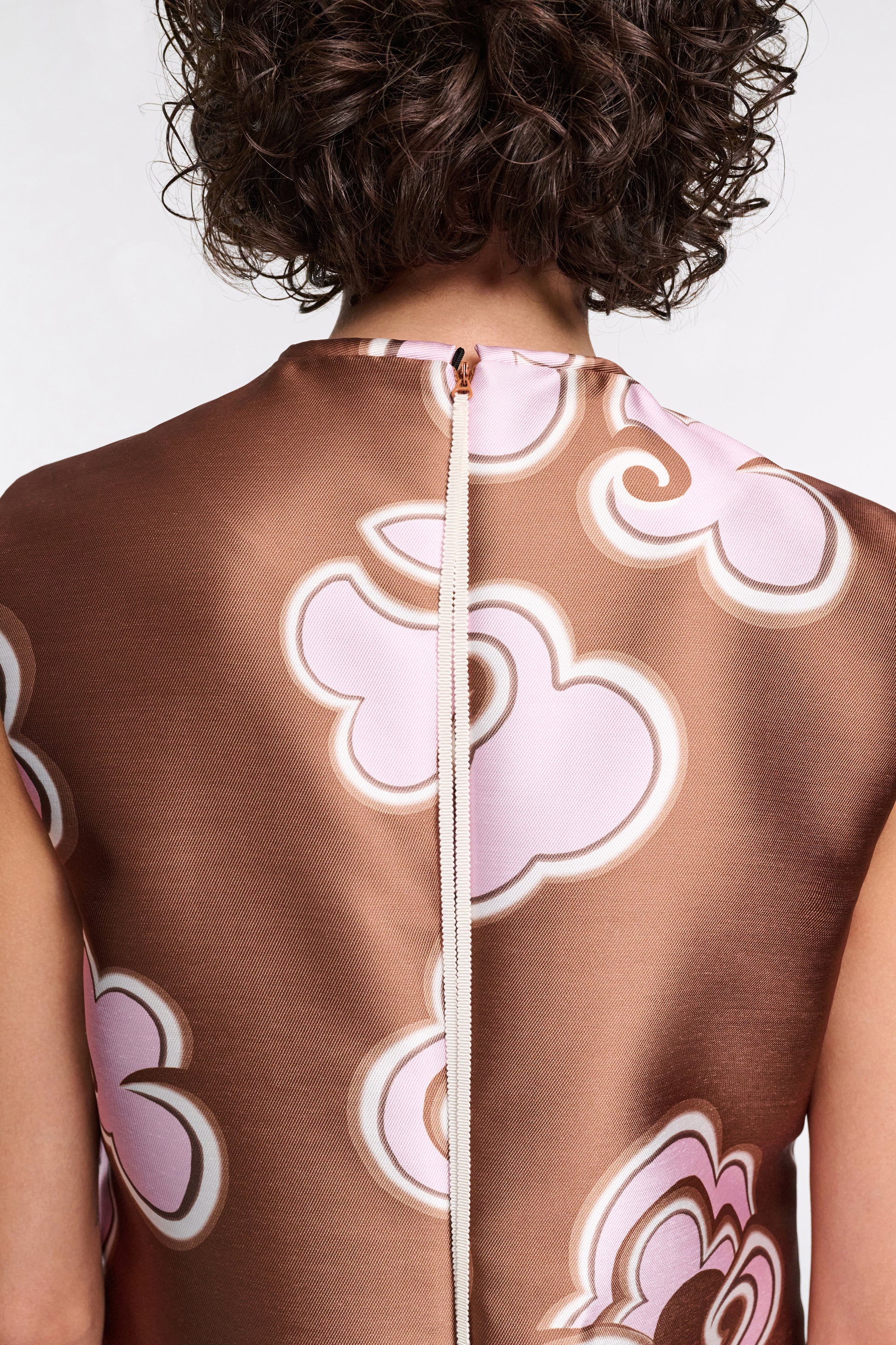 A person with short curly hair is shown from behind wearing the Dorothee Schumacher Floral Statement Dress in brown satin with pink and white cloud patterns, crafted from luxurious duchesse twill and featuring a visible center-back zipper.