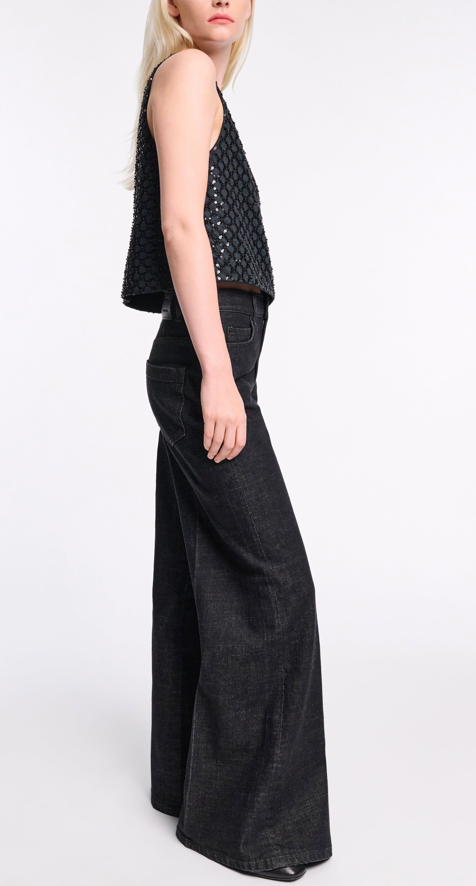 A woman stands in profile against a white background, wearing dark wash, wide leg Deep Love Pants by Dorothee Schumacher and a sleeveless black textured top for a modern, chic look.