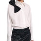 A person wears the Dorothee Schumacher Poplin Power Bow Blouse, a white cotton poplin shirt with an oversized black bow on the shoulder, paired with a black textured mini skirt.