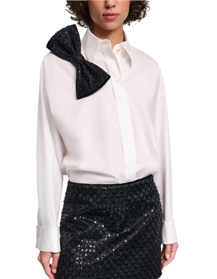 A person wears the Dorothee Schumacher Poplin Power Bow Blouse, a white cotton poplin shirt with an oversized black bow on the shoulder, paired with a black textured mini skirt.