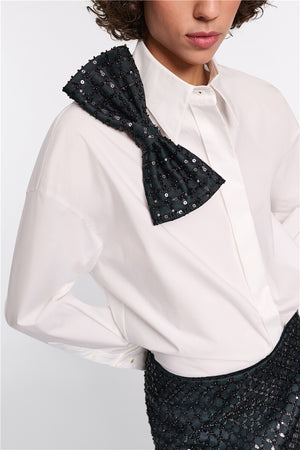 A person wears the Dorothee Schumacher Poplin Power Bow Blouse, an oversized cotton shirt with a large black sequined shoulder bow and delicate sequin floral embroidery, paired with a matching sequined skirt.