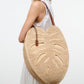 A woman in a white sleeveless dress carries the Staud Large Palm Raffia Bag, a woven straw accessory shaped like a monstera leaf and accented with brown leather handles.