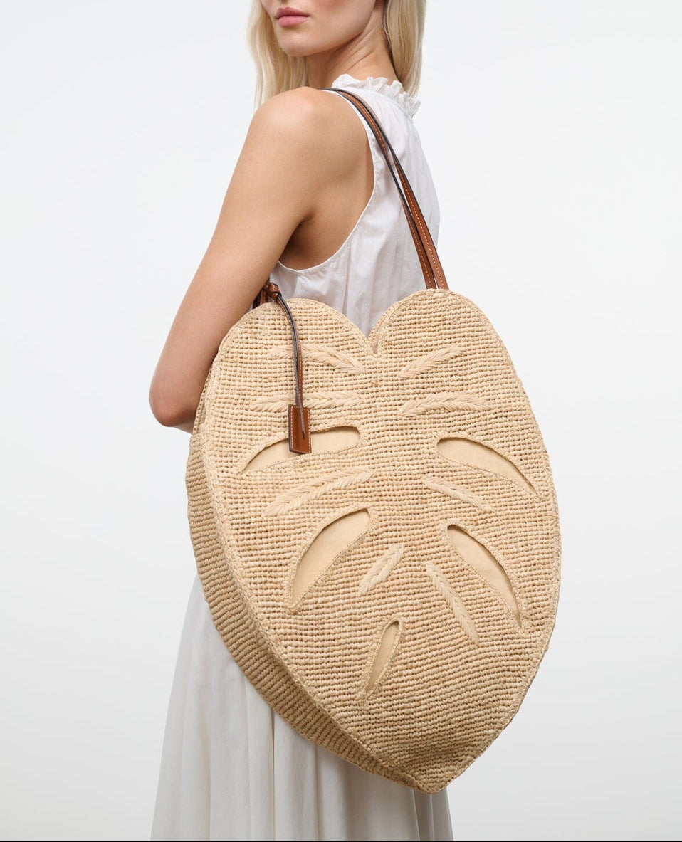 A woman in a white sleeveless dress carries the Staud Large Palm Raffia Bag, a woven straw accessory shaped like a monstera leaf and accented with brown leather handles.