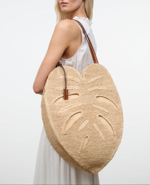 A woman in a white sleeveless dress carries the Staud Large Palm Raffia Bag, a woven straw accessory shaped like a monstera leaf and accented with brown leather handles.