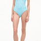 A woman in the Staud Adelaide One Piece—a light blue swimsuit with vertical black stripes and supportive shoulder straps—pairs it with black flip-flops, standing against a plain white background.