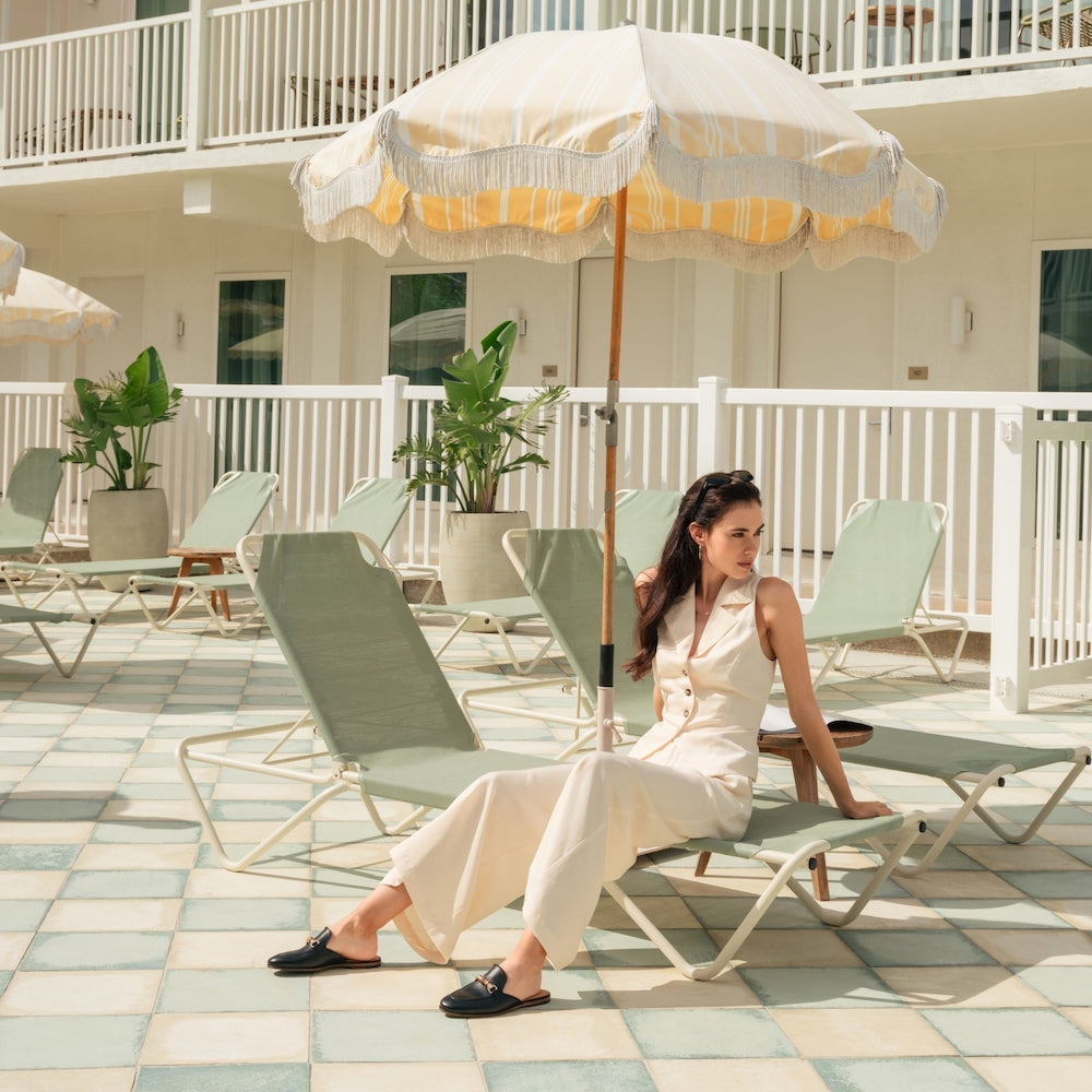 A woman in a light jumpsuit and Magnanni Sienna Luck II Mule loafers relaxes on a lounge chair under a striped umbrella by the poolside, with empty chairs and potted plants in the background.