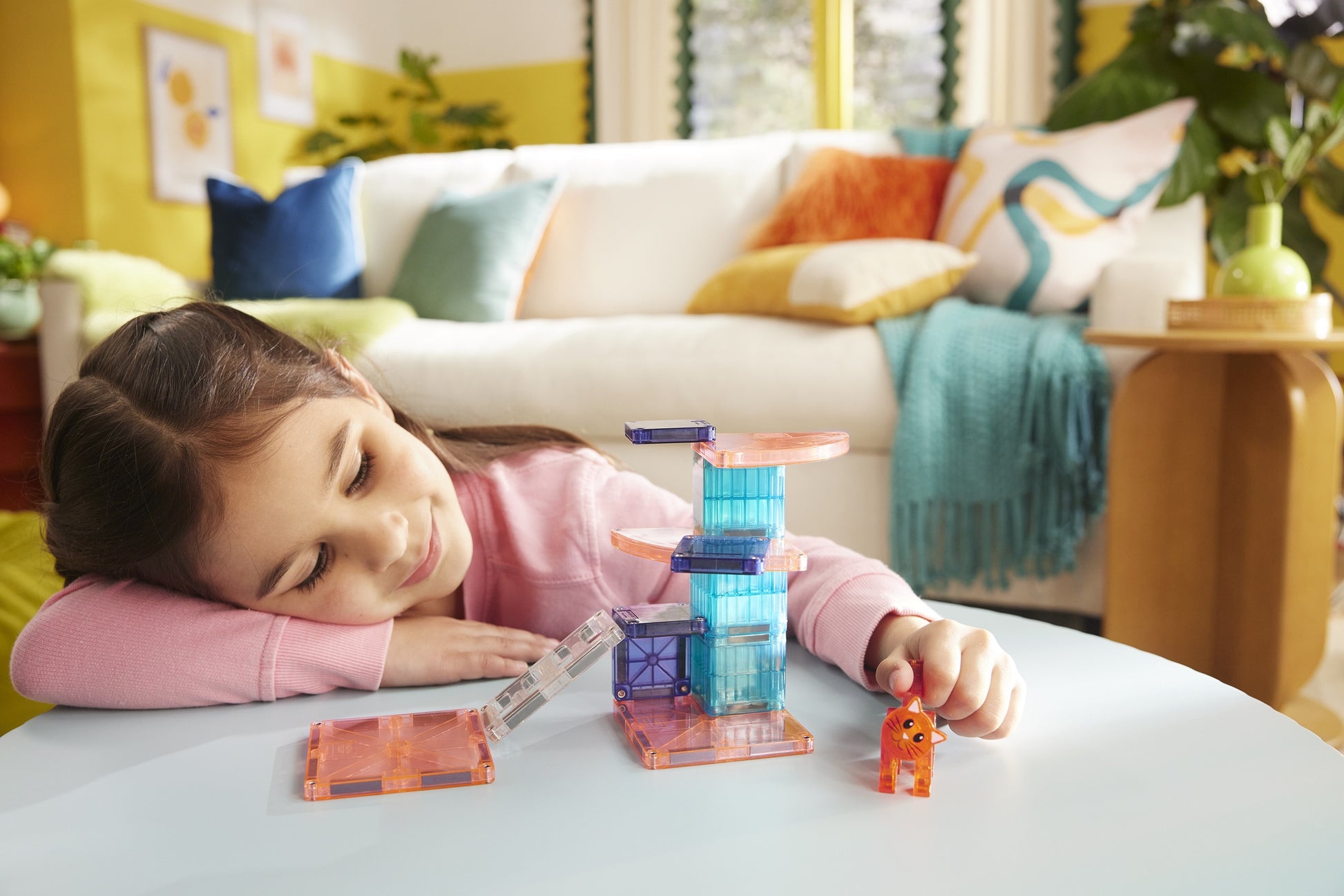 A young girl smiles as she enjoys creative hands-on play with the Magnatiles Cat Tree 13-Piece Set, building and exploring with magnetic pieces and an orange cat figurine in a colorful living room.