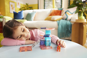 A young girl smiles as she enjoys creative hands-on play with the Magnatiles Cat Tree 13-Piece Set, building and exploring with magnetic pieces and an orange cat figurine in a colorful living room.