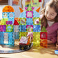 A young girl plays with the Magnatiles City Center 110-Piece Set and toy cars on a track at a table. Colorful structures from her creative Magnatiles construction set and various other playful items are visible around her.