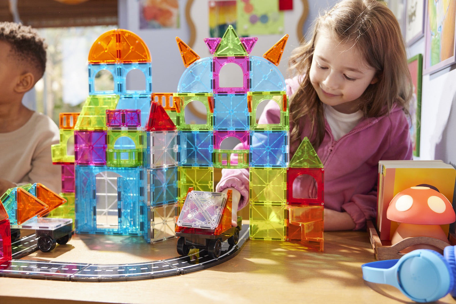 A young girl plays with the Magnatiles City Center 110-Piece Set and toy cars on a track at a table. Colorful structures from her creative Magnatiles construction set and various other playful items are visible around her.