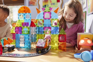 A young girl plays with the Magnatiles City Center 110-Piece Set and toy cars on a track at a table. Colorful structures from her creative Magnatiles construction set and various other playful items are visible around her.