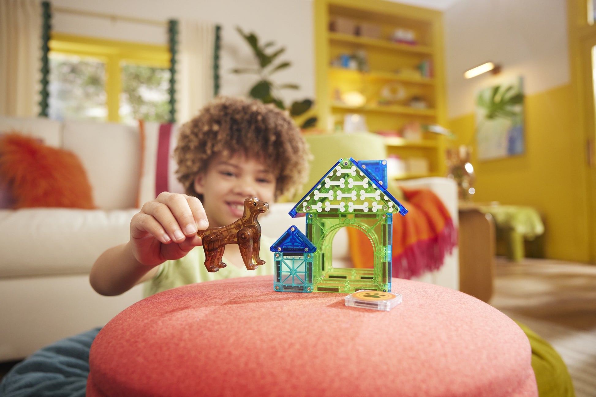 A child plays with the MagnaTiles Dog House 13-Piece Set, featuring colorful pieces, on a red ottoman in a brightly lit living room.