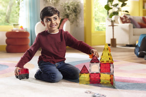 A young boy sits on the floor, smiling as he plays imaginatively with the Magnatiles Fire Rescue 27-Piece Set and a toy car in a brightly lit living room.