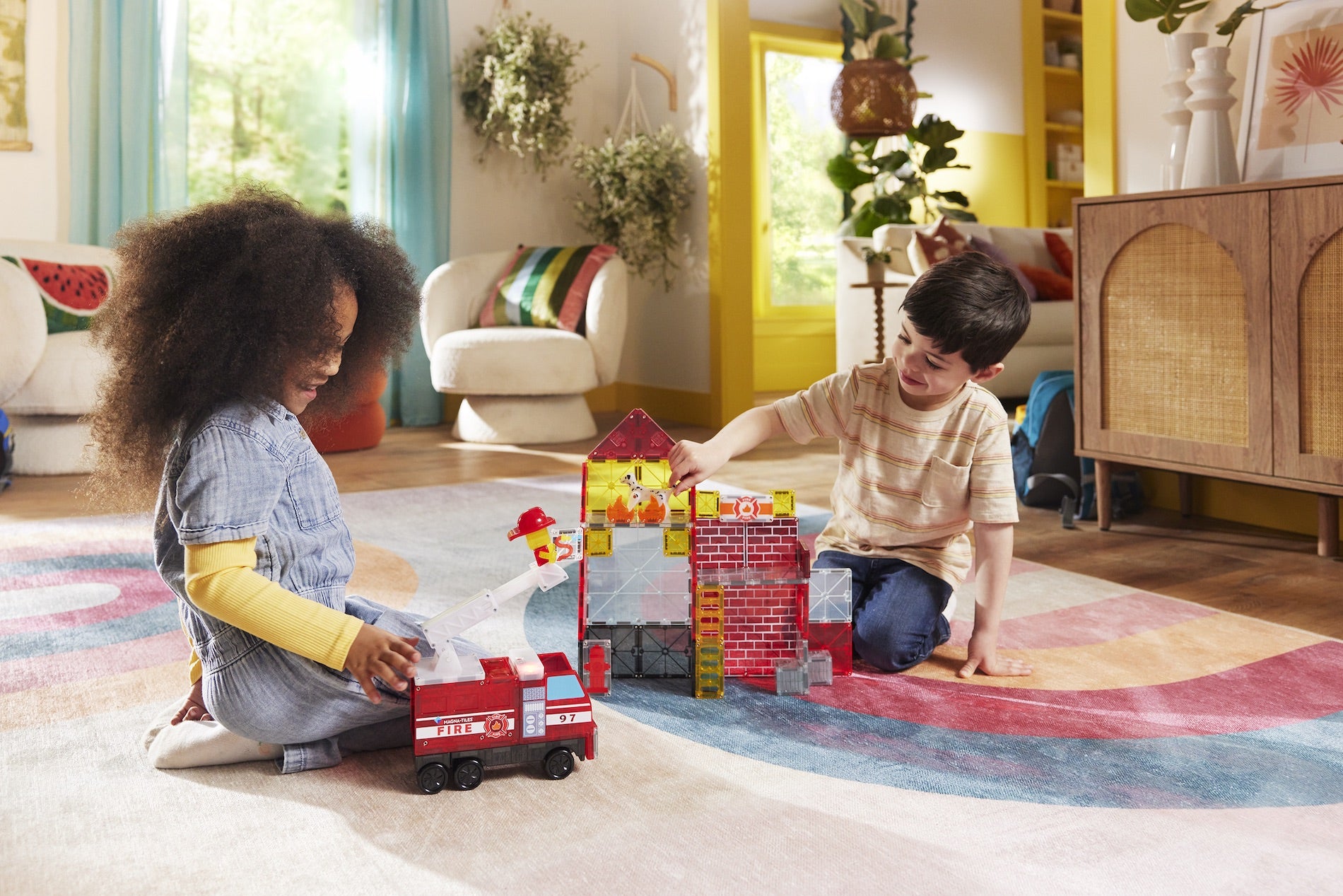Two children sit on a colorful rug, engaging in imaginative play with building blocks, a toy fire truck, and the Magnatiles Fire Station 50-Piece Set in a sunlit living room decorated with plants.