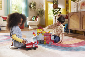 Two children sit on a colorful rug, engaging in imaginative play with building blocks, a toy fire truck, and the Magnatiles Fire Station 50-Piece Set in a sunlit living room decorated with plants.