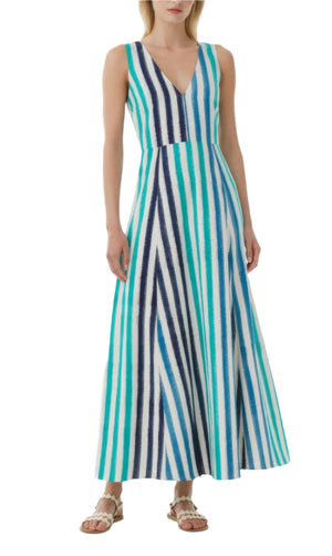 A woman wears the Emporio Sirenuse Nellie Ikat Stripes Dress, a sleeveless, ankle-length style in blue, teal, and white stripes inspired by the ocean, paired with white sandals.