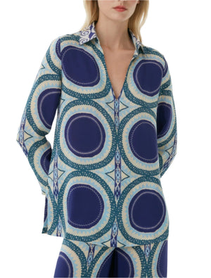 A woman wears the Emporio Sirenuse Jia Solaris Top, a relaxed-fit, long-sleeve V-neck blouse with a pointed collar and blue and teal Solaris print inspired by Suzani textiles.