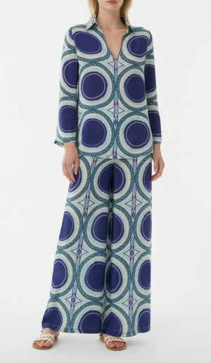 A woman wears the Emporio Sirenuse Jia Solaris Top, featuring a bold blue, green, and cream circular print. She pairs it with white sandals for a stylish look.