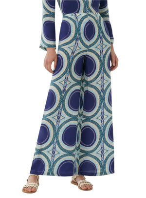 A person wearing Emporio Sirenuse’s Stephan Silk Solaris Pants, featuring a blue and green circular geometric Solaris print, styled with white sandals.