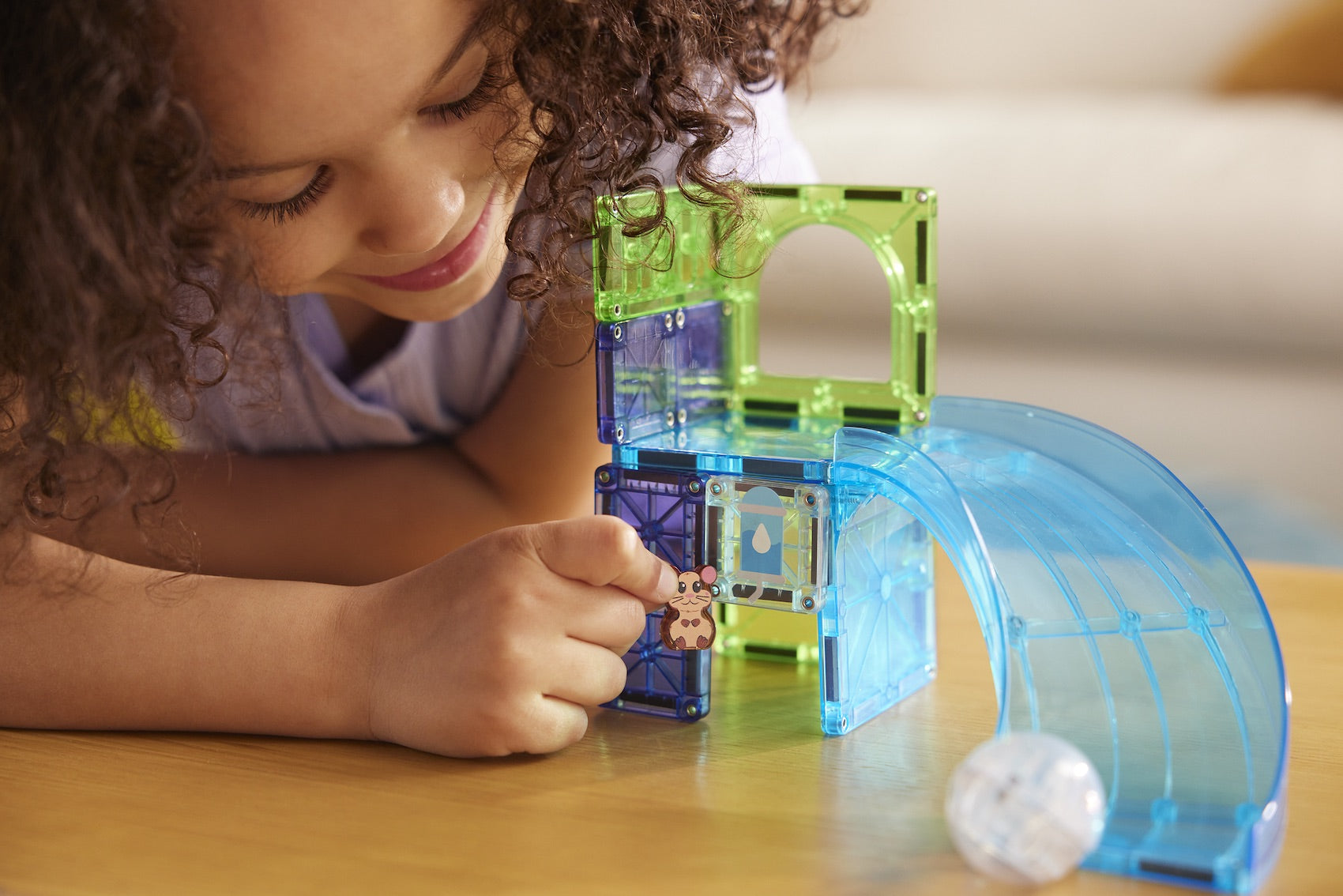 A child sits at a wooden table and plays with the Magnatiles Hamster Ball 13-Piece Set, placing a small animal figure inside the translucent magnetic building pieces.