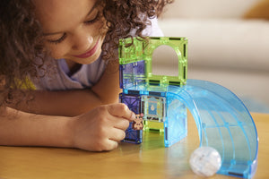 A child sits at a wooden table and plays with the Magnatiles Hamster Ball 13-Piece Set, placing a small animal figure inside the translucent magnetic building pieces.