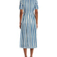 A person in an Emporio Sirenuse Claudia Ikat Stripe Dress, featuring a blue and white pleated stripe design, stands with their back to the viewer while wearing flat sandals.