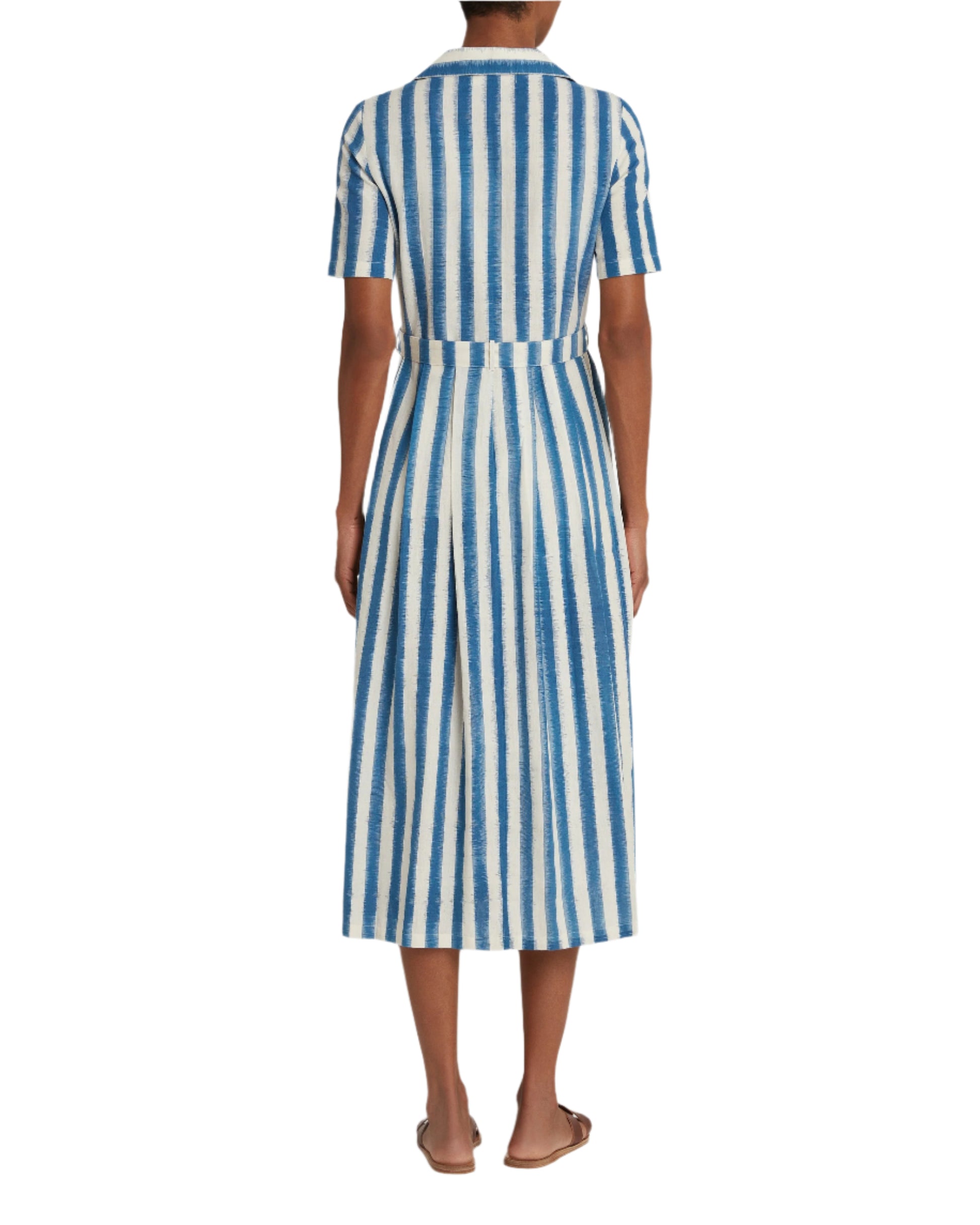 A person in an Emporio Sirenuse Claudia Ikat Stripe Dress, featuring a blue and white pleated stripe design, stands with their back to the viewer while wearing flat sandals.