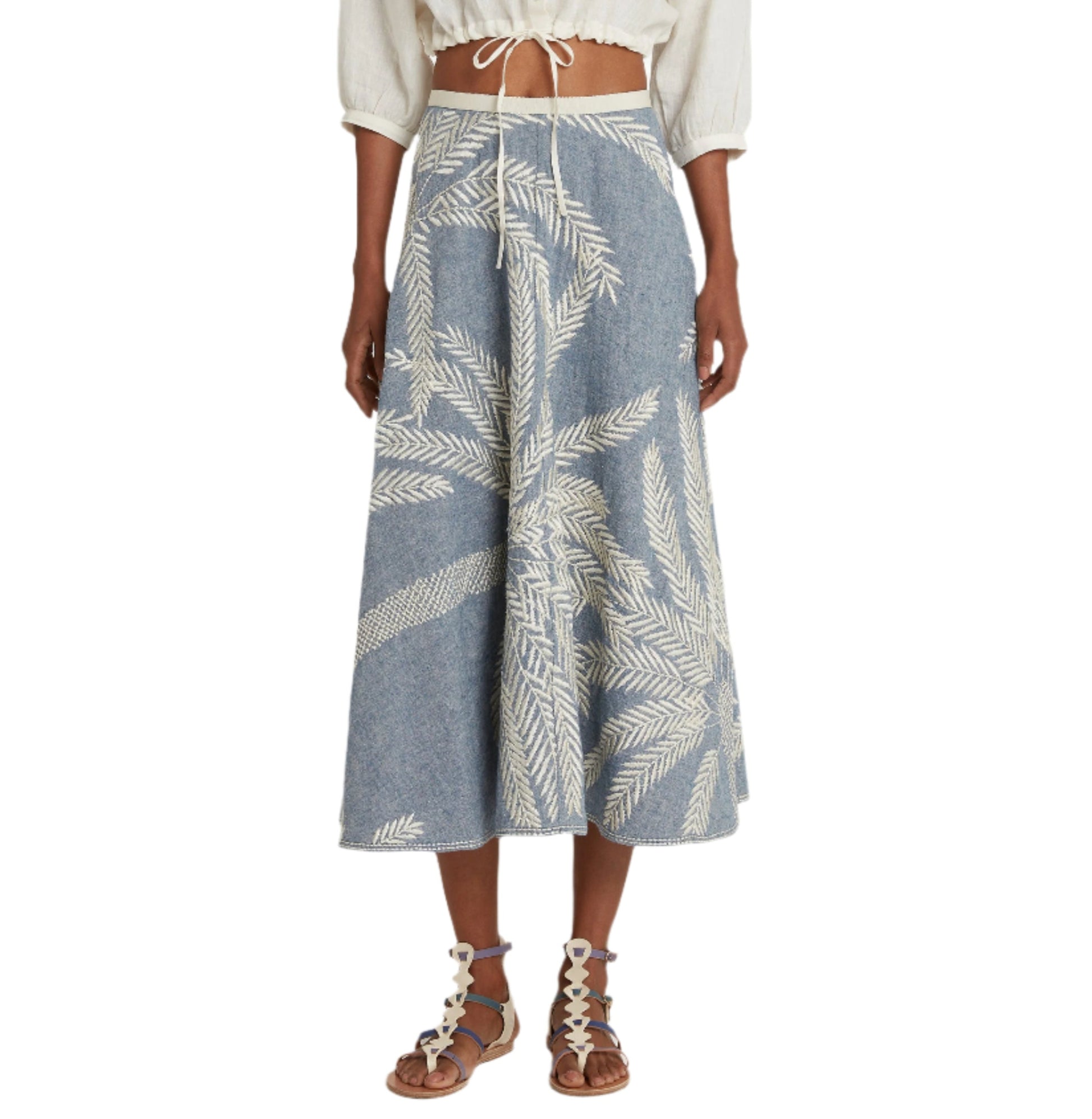 A person wears a white cropped blouse with the Emporio Sirenuse Tasha Palm Embroidery Skirt, featuring white leaf patterns on blue Japanese linen. The look is completed with lace-up sandals, highlighting the intricate palm stitch embroidery.