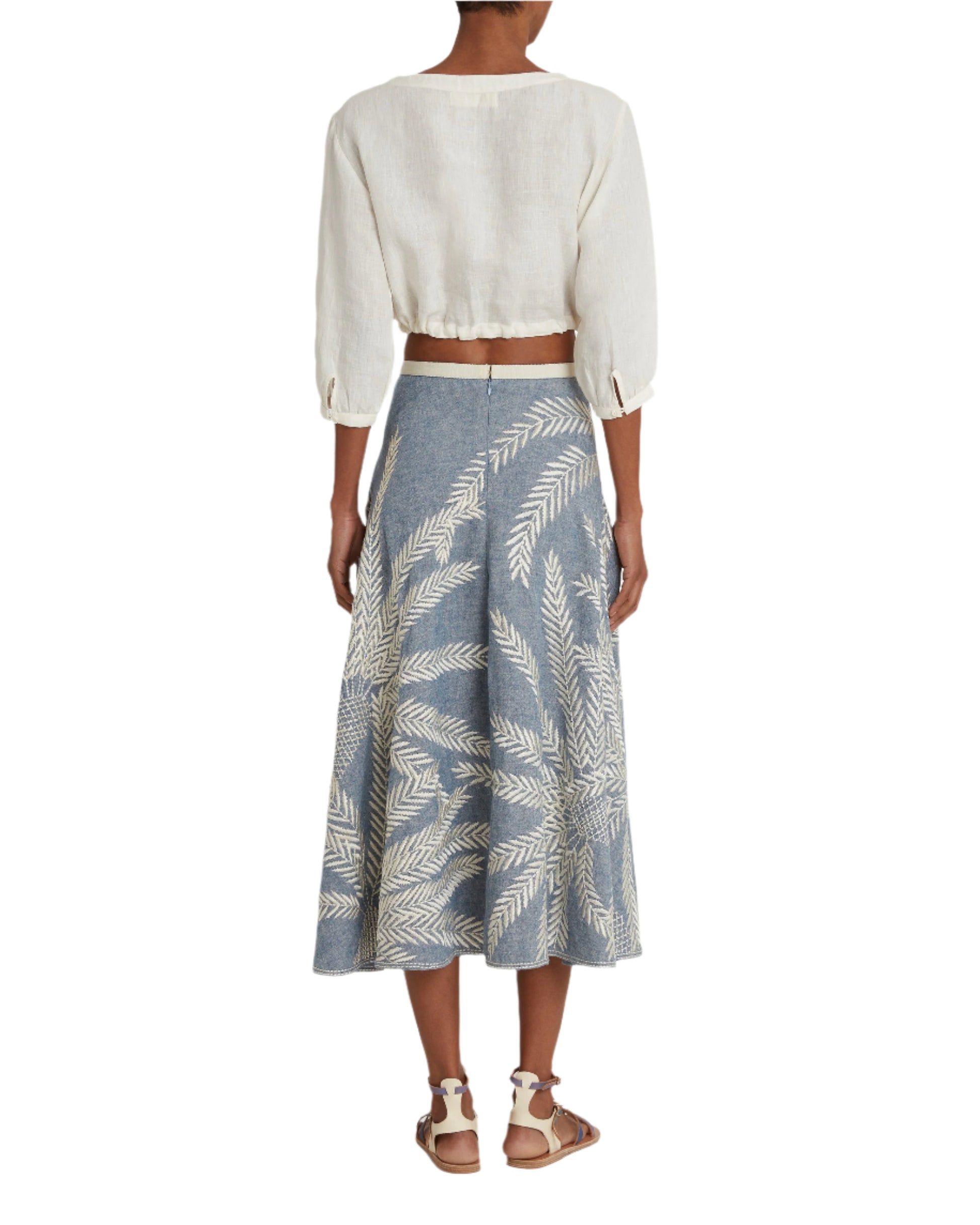 A person is shown from the back wearing a white cropped blouse with the elegant Emporio Sirenuse Tasha Palm Embroidery Skirt in Japanese linen featuring white leaf patterns, paired with white sandals.