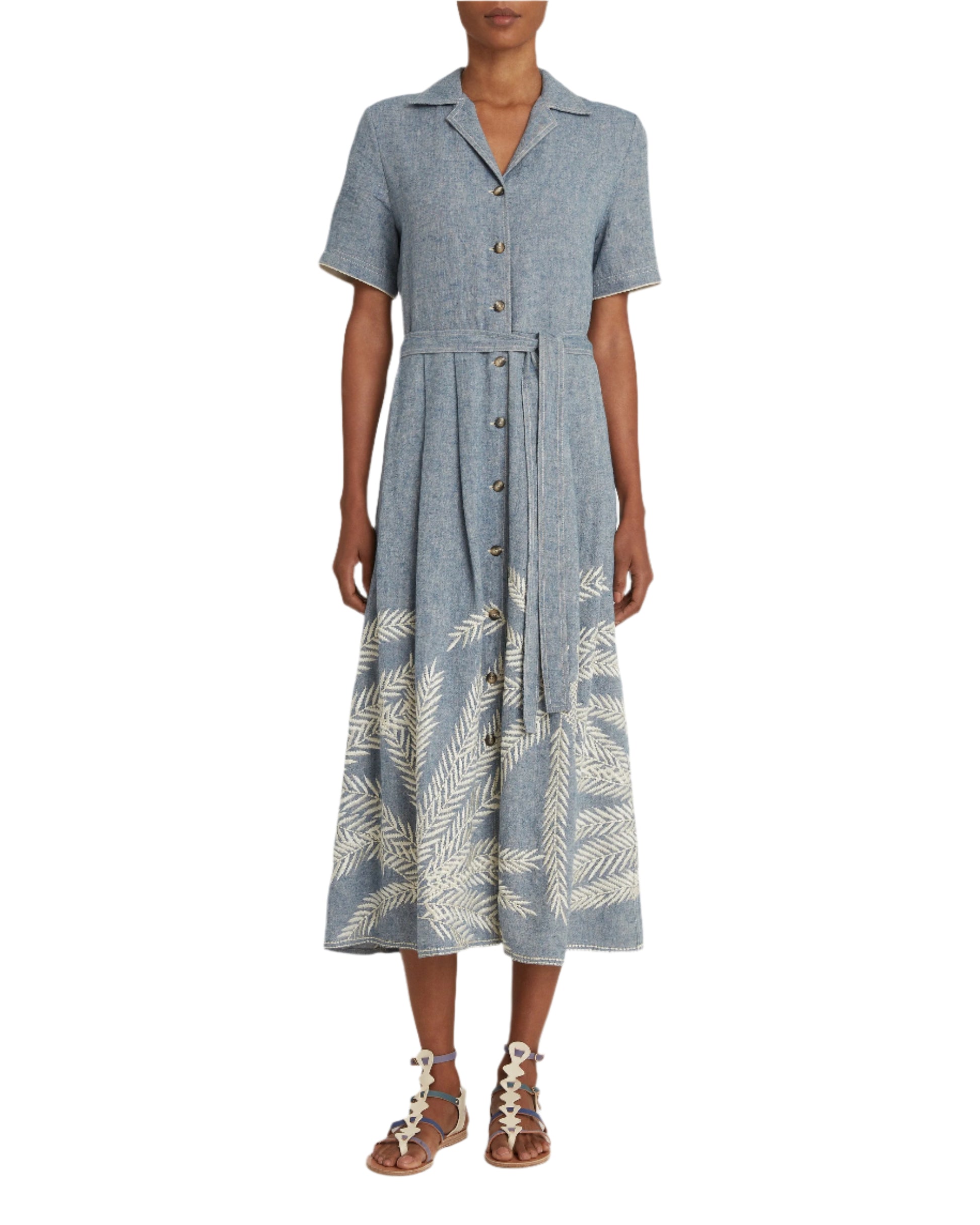 A woman is wearing the Emporio Sirenuse Claudia Palm Embroidery Dress, a blue Japanese linen button-up with a waist tie and white leaf pattern near the hem, paired with strappy sandals.