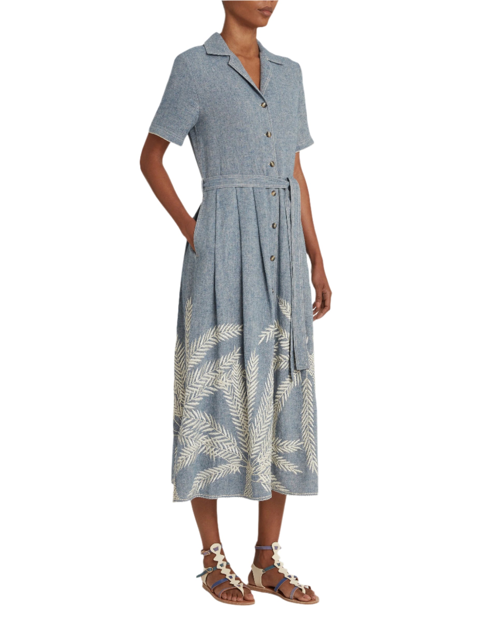 An individual wears an Emporio Sirenuse Claudia Palm Embroidery Dress in blue with fern patterns on the hem, made from elegant Japanese linen, paired with white sandals.