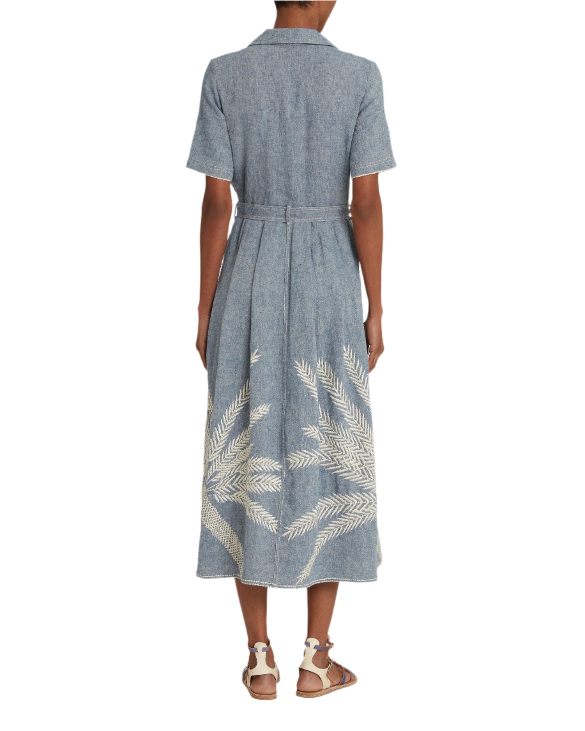 Back view of a person in an Emporio Sirenuse Claudia Palm Embroidery Dress, showcasing white palm-stitched designs on blue chambray Japanese linen, with short sleeves, a collar, and waist tie, paired with tan sandals.