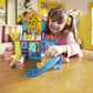 A young girl smiles while playing with colorful Magnatiles, including the Pet Playhouse 50-Piece Set, and toy figures on a wooden table in a bright living room.