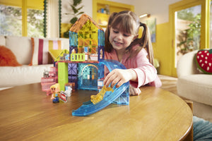 A young girl smiles while playing with colorful Magnatiles, including the Pet Playhouse 50-Piece Set, and toy figures on a wooden table in a bright living room.