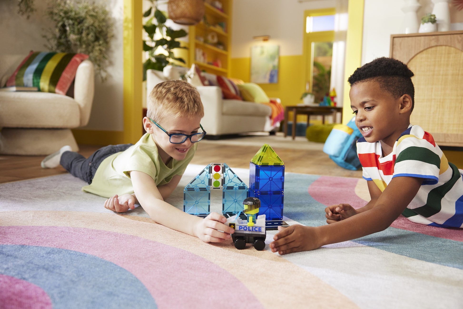 Two children play in a bright living room, building with the Magna-Tiles Police Station 20-Piece Set and racing the included toy police car on a colorful rug.