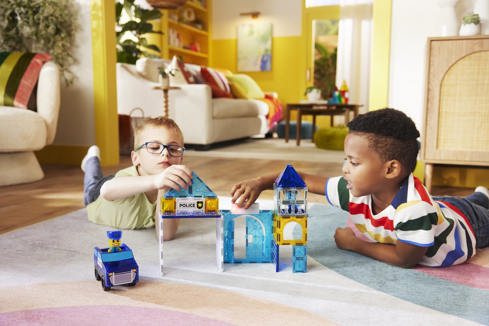 Two boys lie on the living room floor, playing with the Magnatiles Police Station 35-Piece Set, which includes a toy police car and a light and sound tile.
