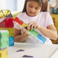 A young girl in a white shirt builds a geometric structure at a white table with the vibrant Magnatiles Rail Racers 90-Piece Set, engaging in hands-on STEM learning.
