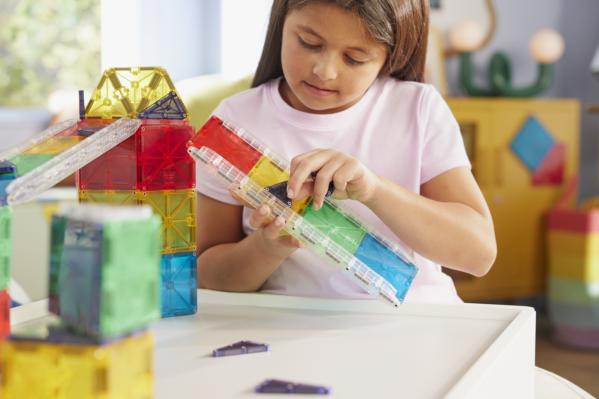 A young girl in a white shirt builds a geometric structure at a white table with the vibrant Magnatiles Rail Racers 90-Piece Set, engaging in hands-on STEM learning.