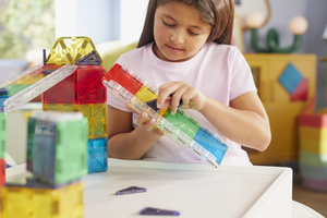 A young girl in a white shirt builds a geometric structure at a white table with the vibrant Magnatiles Rail Racers 90-Piece Set, engaging in hands-on STEM learning.