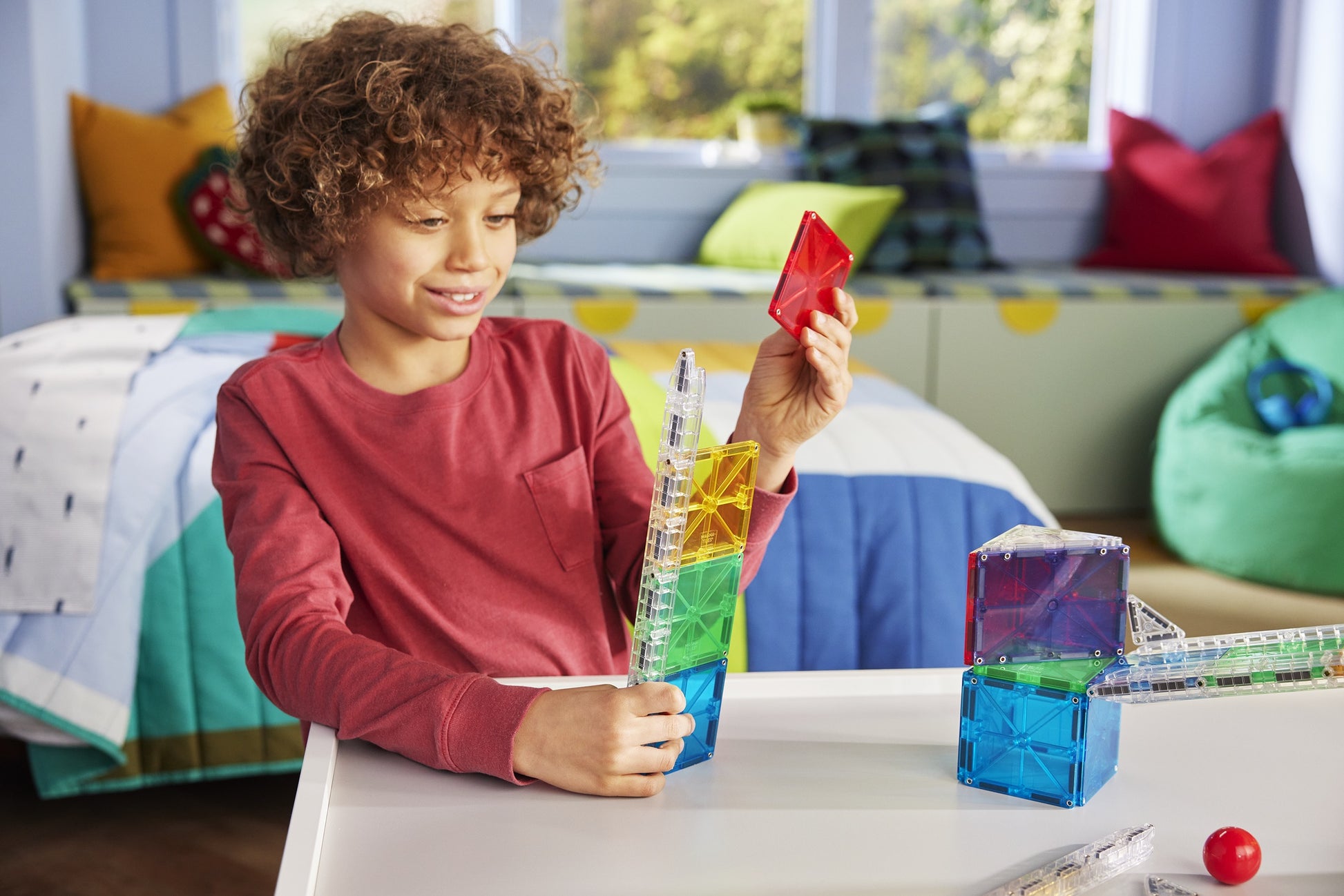 A child smiles while building a colorful structure with the Magnatiles Rail Racers 33-Piece Set, a STEM toy, in their bedroom.