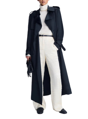 Wearing the Simkhai Alvida Belted Long Coat over a white turtleneck and textured white pants, this look is finished with a black belt, black shoes, and a small black handbag. The ensemble showcases the timeless elegance of the double-breasted coat by Simkhai, elevated with chic accessories for a polished appearance.