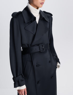 Dressed in the Simkhai Alvida Belted Long Coat—a stylish black, double-breasted piece with a defined waist—layered over a white turtleneck sweater, the individual stands against a plain light background.