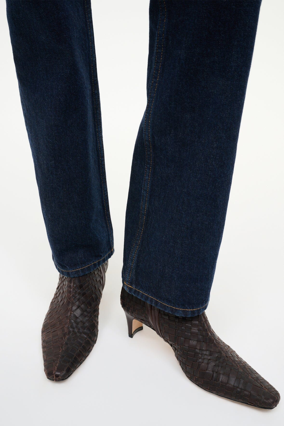 Person wearing dark blue jeans and Staud Wally Woven Leather Ankle Boots with low blade heels, standing on a light background.
