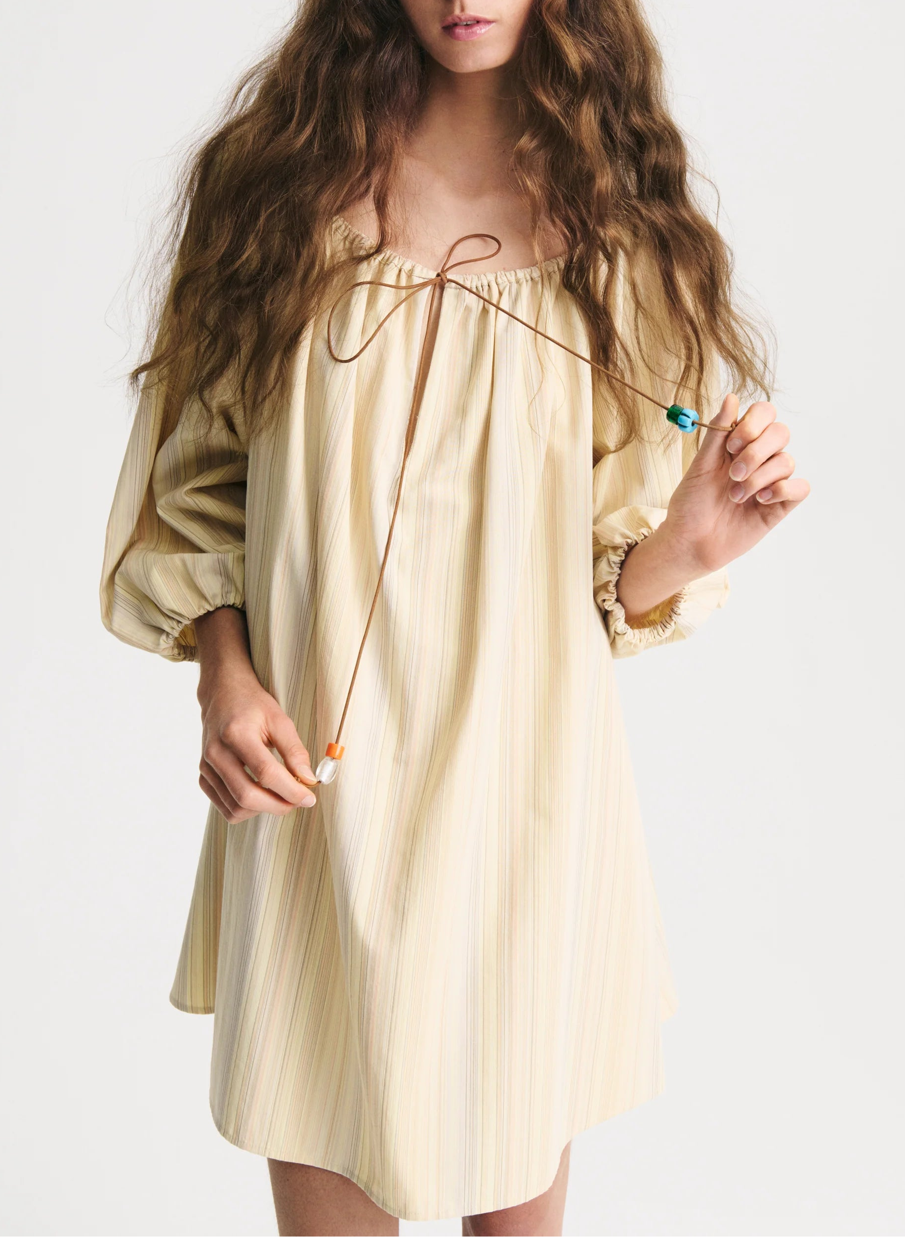 A woman with long, wavy hair wears the Bernadette Bonnie Short Stripe Dress—a loose summer style with puffed sleeves and a tie neckline—holding the ties in both hands.