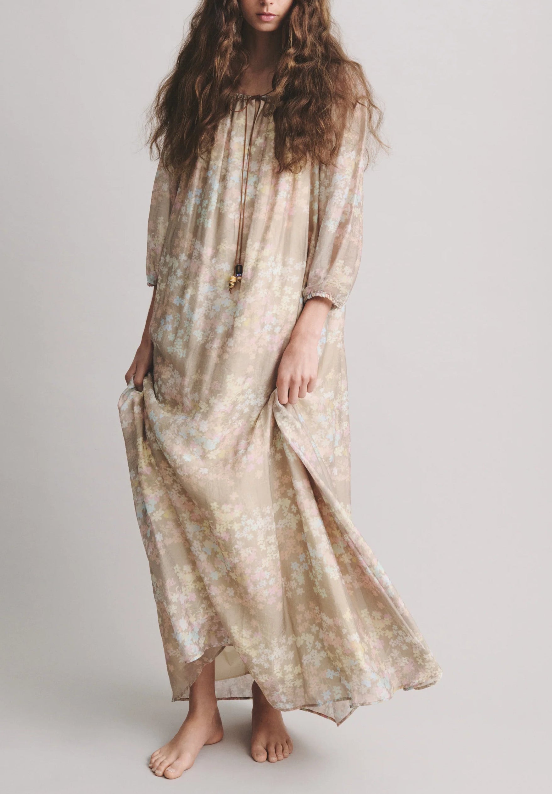 A person with long wavy hair stands barefoot, holding one side of their loose, light beige Bernadette Bonnie Maxi Jasmin Dress—a perfect summer dress.