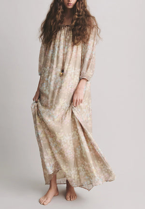 A person with long wavy hair stands barefoot, holding one side of their loose, light beige Bernadette Bonnie Maxi Jasmin Dress—a perfect summer dress.