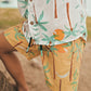 A child stands barefoot outdoors in Pepita & Me—wearing a white shirt and Boys' Swim Trunks, both featuring green palm trees and orange suns on quick-dry fabric.
